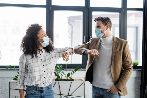 Young interracial business colleagues in medical masks doing elbow bump in office — Stock Photo