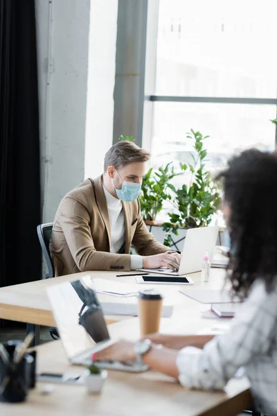 African american manager typing on laptop near colleague in medical mask, blurred foreground — Stock Photo