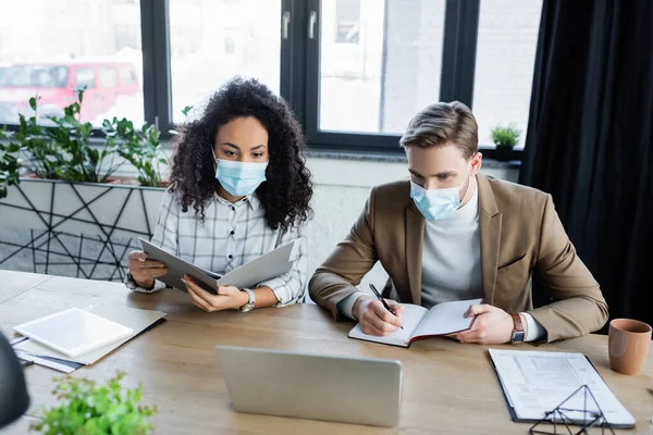 Interracial business colleagues in medical masks working near laptop with notebook and documents — Stock Photo