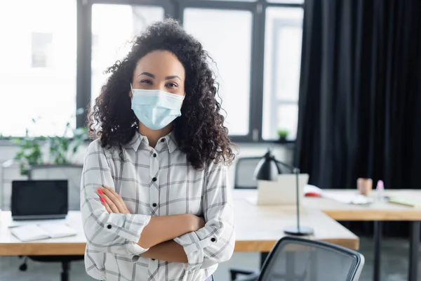 Femme d'affaires afro-américaine positive en masque médical debout avec les bras croisés au bureau — Photo de stock