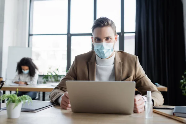 Молодий менеджер в медичній масці дивиться на камеру біля ноутбука і афроамериканського колеги на розмитому фоні — стокове фото