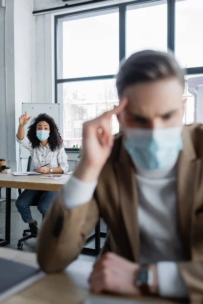 Mujer de negocios afroamericana en máscara protectora agitando la mano detrás de colega que sufre de dolor de cabeza en primer plano borrosa - foto de stock