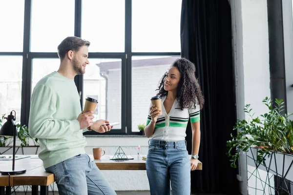 Happy interracial business partners standing with coffee to go during conversation in office — Stock Photo