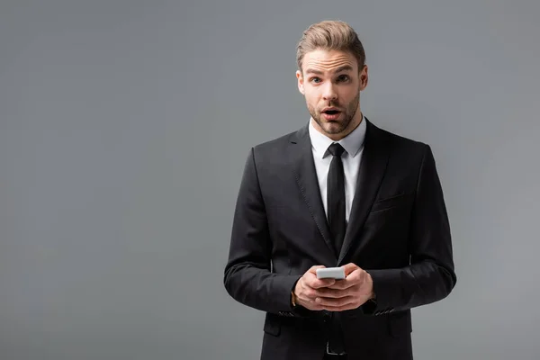 Шокированный бизнесмен, смотрящий в камеру во время чата на смартфоне, изолированном от серого — стоковое фото