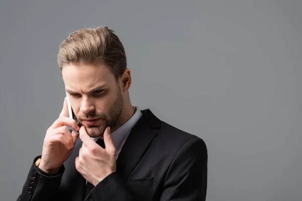 Empresario pensativo tocando la barbilla mientras habla en el teléfono celular aislado en gris - foto de stock