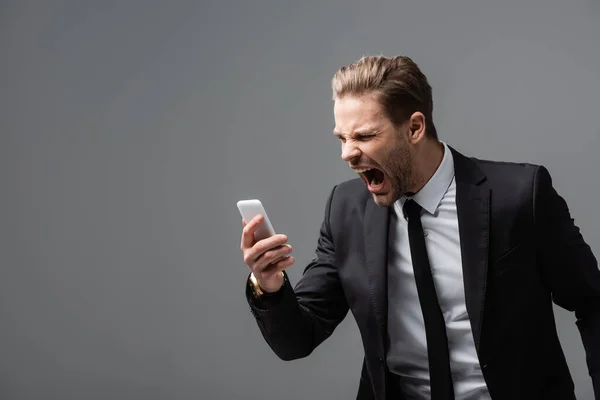 Разъяренный бизнесмен кричит во время видеозвонка на мобильный телефон, изолированный на серый — стоковое фото