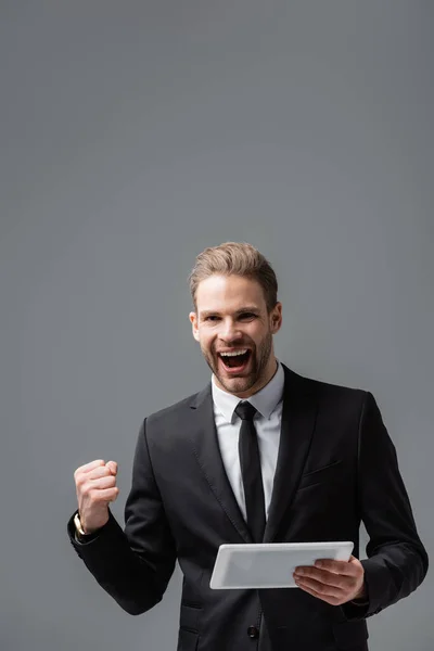 Excited businessman showing success gesture while holding digital tablet isolated on grey — Stock Photo