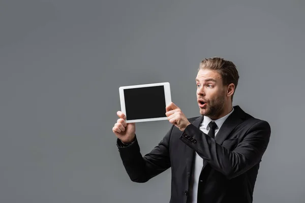Шокированный бизнесмен с открытым ртом показывает цифровой планшет с чистым экраном изолированы на сером — стоковое фото