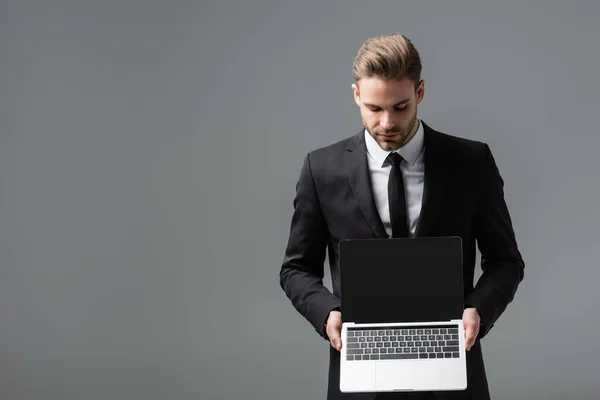 Young businessman in black suit holding laptop with blank screen isolated on grey — Stock Photo