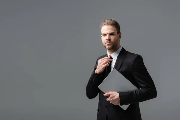 Serious businessman holding laptop while adjusting tie isolated on grey — Stock Photo