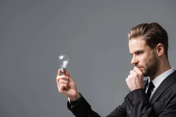 Thinking businessman holding hand near face while looking at light bulb isolated on grey — Stock Photo