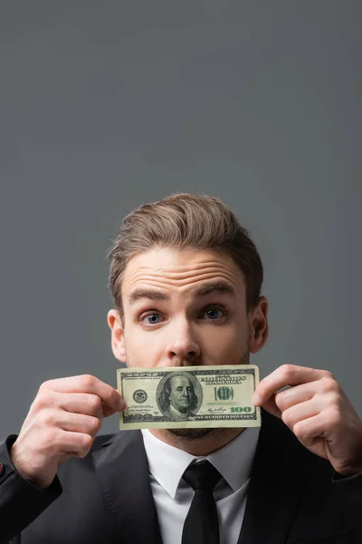 Excited businessman covering mouth with dollar banknote isolated on grey — Stock Photo