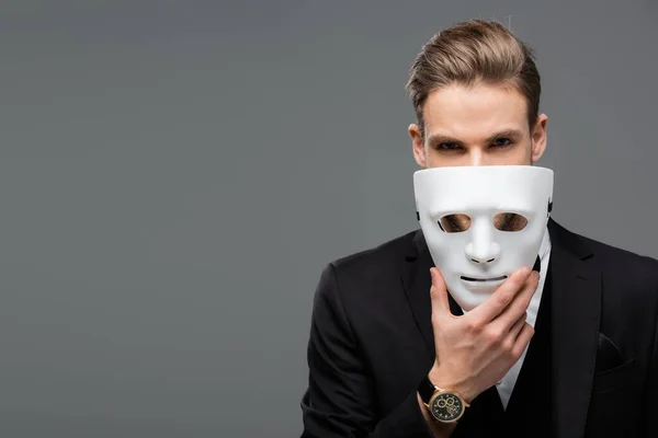 Hombre de negocios sosteniendo la máscara cerca de la cara mientras mira la cámara aislada en gris - foto de stock