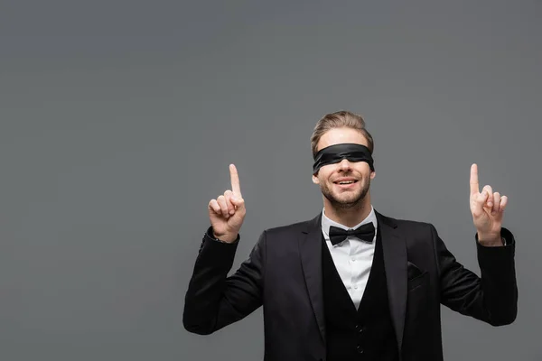 Smiling businessman in blindfold pointing up with fingers isolated on grey — Stock Photo