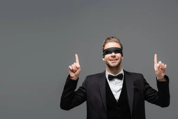 Hombre de negocios sonriente con los ojos vendados apuntando hacia arriba con los dedos aislados en gris - foto de stock