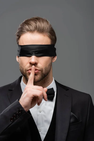 Joven hombre de negocios con los ojos vendados mostrando un signo de silencio aislado en gris - foto de stock