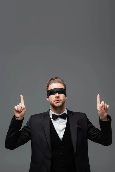 Joven hombre de negocios con los ojos vendados apuntando hacia arriba con los dedos aislados en gris - foto de stock