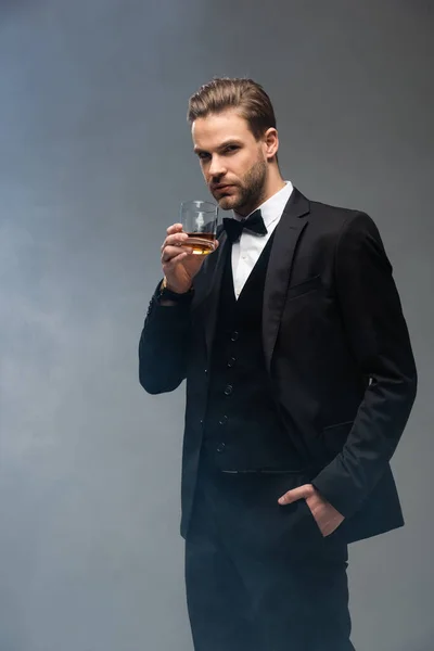 Elegant businessman standing with hand in pocket while holding whiskey on grey background with smoke — Stock Photo