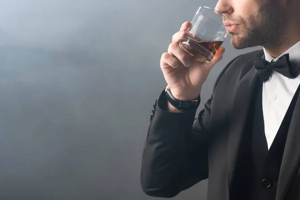 Vista parcial del elegante hombre de negocios bebiendo whisky sobre fondo gris con humo - foto de stock