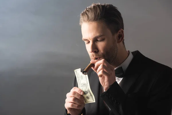 Young businessman lighting cigar with hundred dollar banknote on grey background with smoke — Stock Photo