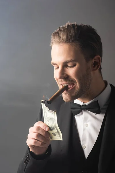 Wealthy businessman lighting cigar with hundred dollar banknote on grey background with smoke — Stock Photo