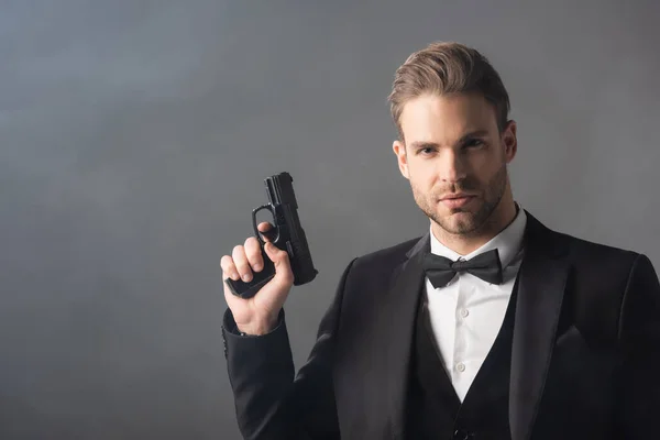 Elegante hombre de negocios mirando a la cámara mientras sostiene el arma sobre fondo gris con humo - foto de stock