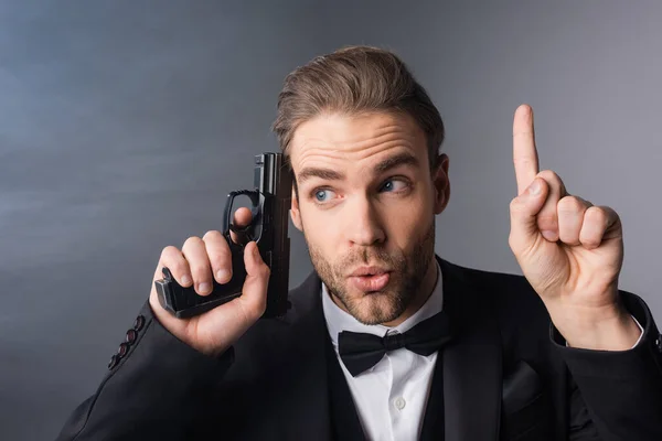 Excitado hombre de negocios mostrando gesto de idea mientras sostiene el arma cerca de la cabeza sobre fondo gris con humo - foto de stock