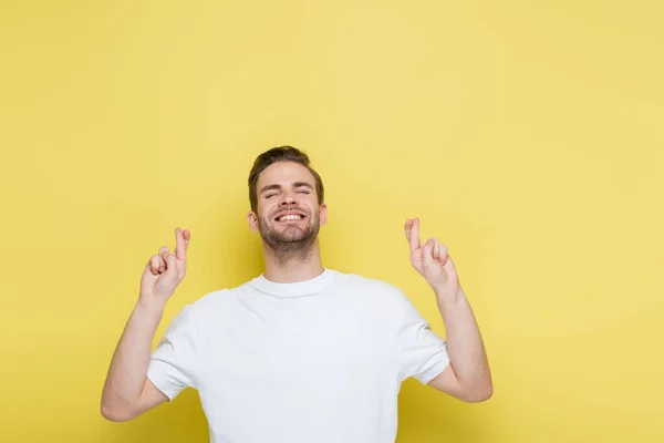 Cheerful man with closed eyes standing with crossed fingers on yellow — Stock Photo