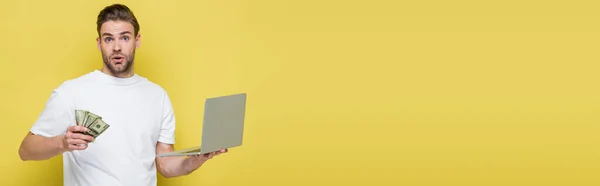 Amazed man looking at camera while holding laptop and money on yellow, banner — Stock Photo