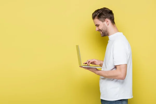 Vista lateral del hombre alegre en camiseta blanca usando el ordenador portátil en amarillo - foto de stock