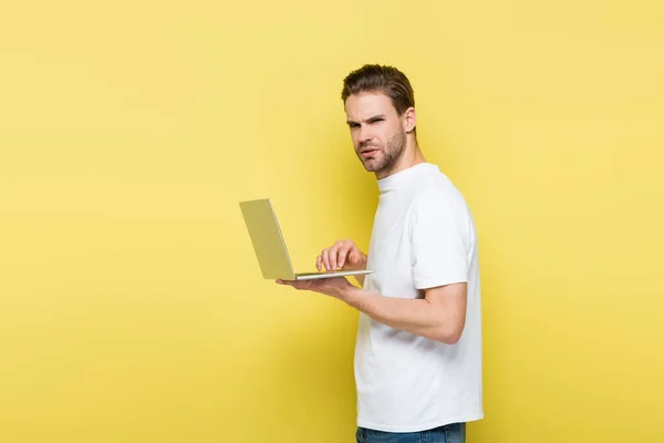 Displeased man looking at camera while typing on laptop on yellow — Stock Photo