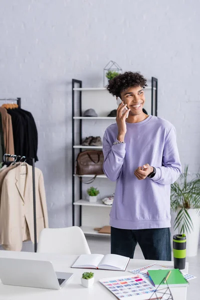 Alegre afroamericano propietario de la sala de exposición hablando en el teléfono celular cerca de la taza termo y portátiles en la mesa - foto de stock