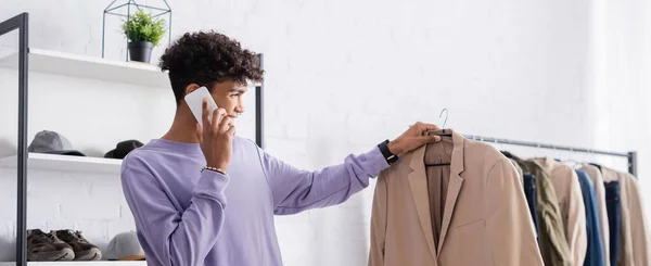 Souriant propriétaire afro-américain de showroom tenant cintre avec veste et parlant sur smartphone, bannière — Photo de stock