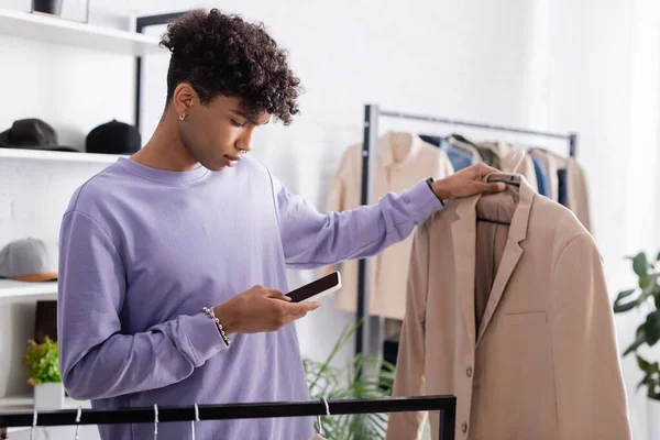 Propriétaire d'un showroom afro-américain utilisant un smartphone et tenant une veste sur un cintre sur fond flou — Photo de stock