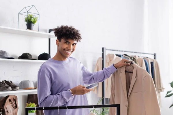 Propriétaire afro-américain de showroom souriant à la caméra tout en tenant smartphone et veste — Photo de stock