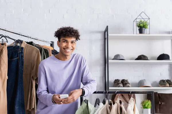 Alegre propietario afroamericano Showroom sosteniendo teléfono inteligente cerca de la ropa - foto de stock