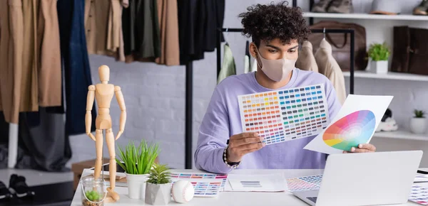 African american showroom owner in protective mask looking at color swatches near laptop, banner — Stock Photo