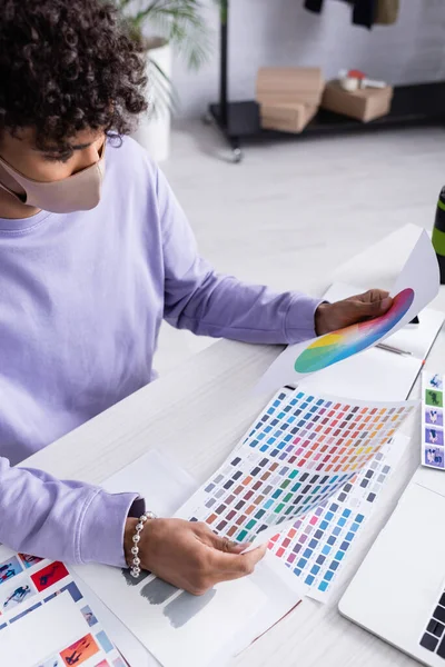 Владелец американского шоу-рума в защитной маске держит красочные образцы рядом с ноутбуком на столе — стоковое фото