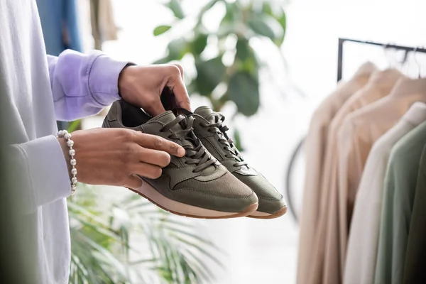 Cropped view of african american owner of showroom holding sneakers near clothes on blurred background — Stock Photo