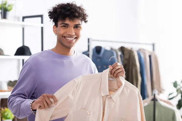 Souriant propriétaire afro-américain de showroom tenant chemise sur cintre et souriant à la caméra — Photo de stock