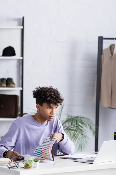 Joven afroamericano propietario de showroom celebración de muestras de colores y mirando a la computadora portátil - foto de stock