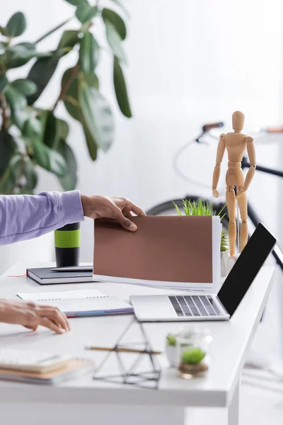 Ausgeschnittene Ansicht eines afrikanisch-amerikanischen Kleinunternehmensbesitzers, der Papiermappe in der Nähe von Laptop mit leerem Bildschirm im Showroom nimmt — Stockfoto