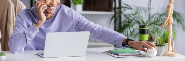 Cropped view of african american showroom owner taking baseball ball while talking on smartphone at working table, banner — Stock Photo