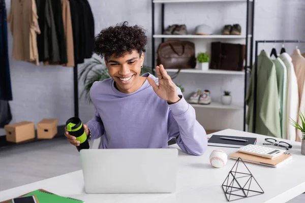 Smiling african american owner of showroom holding thermo cup during video call on laptop — Stock Photo