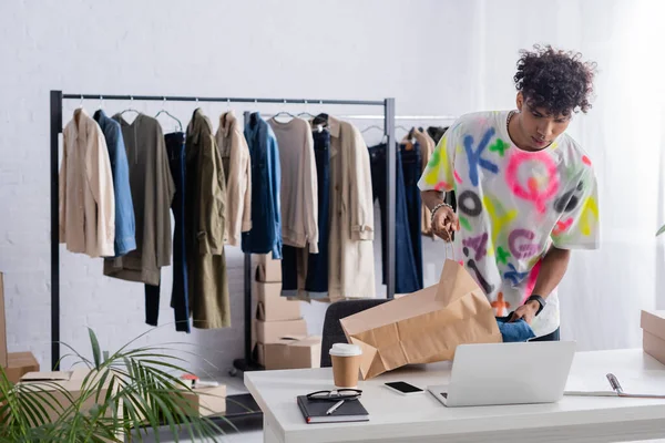 Африканский американец владелец выставочного зала положить одежду в торговый мешок рядом с устройствами и кофе, чтобы пойти — стоковое фото