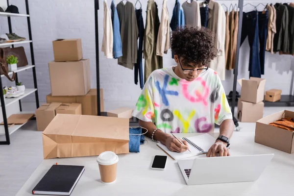 Стильный африканский американец владелец шоурума, использующий ноутбук и пишущий на ноутбуке рядом с пакетами и кофе, чтобы пойти — стоковое фото