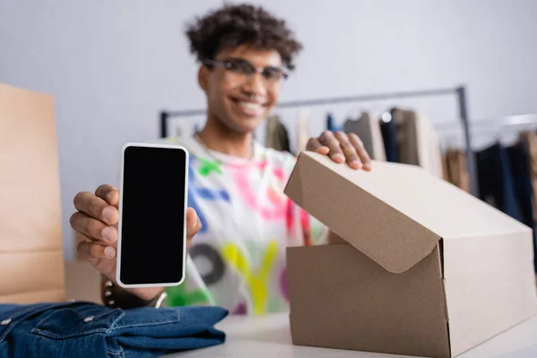 Смартфон з чистим екраном у руці афроамериканського бізнесмена на розмитому фоні біля одягу та коробки. — стокове фото
