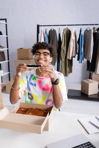 Усміхнений афроамериканський власник шоуруму фотографує на смартфоні біля коробки з одягом і блокнотом — стокове фото