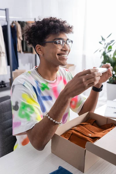 Joyeux propriétaire afro-américain de showroom prenant des photos sur smartphone près de la boîte en carton avec des vêtements — Photo de stock