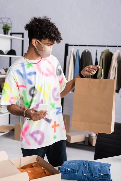 African american showroom owner in protective mask holding smartphone and shopping bag — Stock Photo